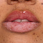 Glossy pink lips exfoliating with a sugar‑crystal lip scrub, bottom lip coated in grainy treatment while the top lip shines with balm, showcasing a ColourPop lip care product in a beauty demo against natural freckled skin.