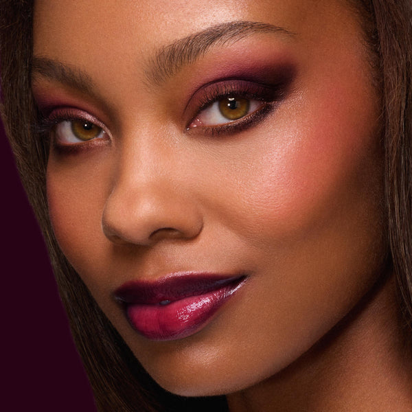 Model wearing rich berry smokey eyeshadow with shimmer and glossy wine lips from A Shadow of Secrets Collection, a ColourPop Full Collection Set, shown in a close-up beauty shot against a deep burgundy background.