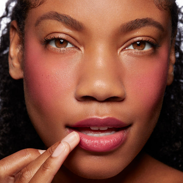 Model finger-applies Girls Night ColourPop Lip and Cheek Balm to lower lip, revealing a glossy berry-pink tint with matching rosy cheeks in a tight, well-lit studio close-up.