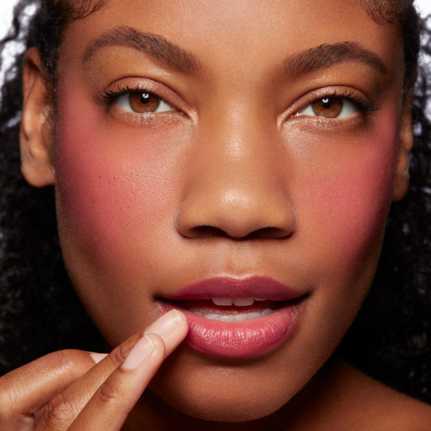 Model finger-applies Girls Night ColourPop Lip and Cheek Balm to lower lip, revealing a glossy berry-pink tint with matching rosy cheeks in a tight, well-lit studio close-up.