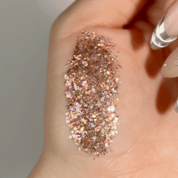 Avenue of the Stars Glitter Gel by ColourPop swatched on a hand, shimmering with multi-size rose-gold and iridescent hex glitter that shifts under light, showing dense sparkle payoff and a smooth, buildable gel texture in a close-up view.