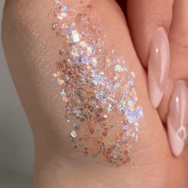 Disco Lady ColourPop Glitter Gel swatched on the back of a hand, shimmering with multicolor iridescent hex and micro-glitter in a clear gel base, catching light beside glossy nude-pink nails in a tight beauty close-up.