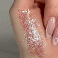 ColourPop Glitter Gel So Cute swatched on the back of a hand, sparkling as chunky and fine iridescent pink hex glitter shifts to blue and silver; glossy, quick-drying gel shows high-shine payoff beside soft pink acrylic nails.