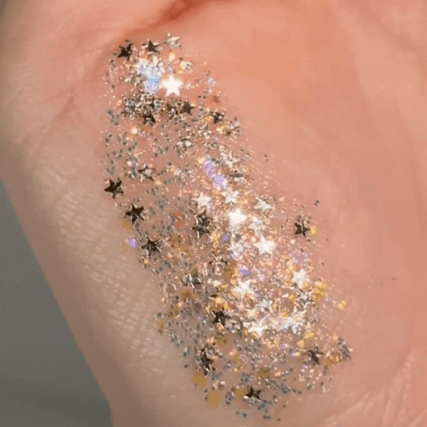 Spinner glitter gel swatched across the back of a hand, flashing holographic rainbow shifts in a clear base with silver microglitter and black star confetti; ColourPop Glitter Gel sparkle and texture shown for face and body looks.