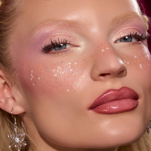 Glossy pink lips wearing ColourPop So Juicy Plumping Gloss Stick in Pinky Who reflecting high shine, framed by soft pink shimmer eyeshadow and star confetti on cheeks, with a dangling snowflake earring in a studio close-up beauty shot.