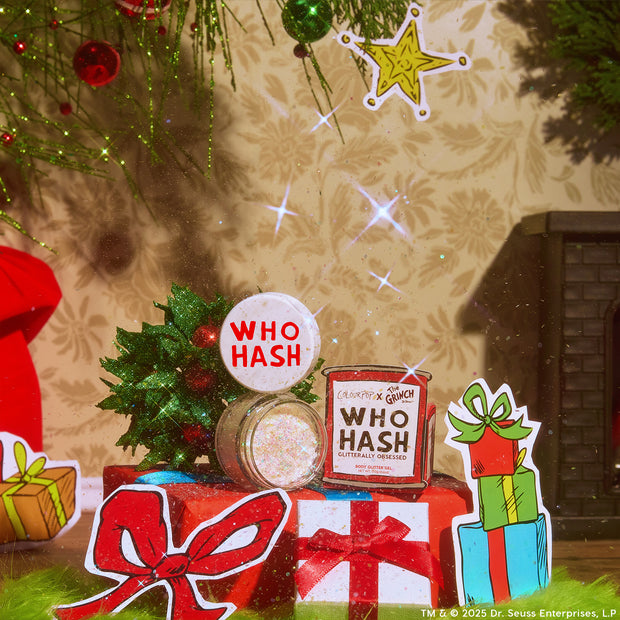 ColourPop Who-Hash glitter gel shines from an open jar with a white lid stamped WHO HASH beside a Grinch-themed tin, displayed among holly, ornaments, and wrapped gifts in a cozy holiday setting, showing iridescent multi-color sparkle.