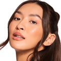 Glossy nude lips wearing Caramel Latte Fresh Kiss Glossy Balm, delivering hydrating shine and a sheer warm caramel tint; full-face close-up of a model with dewy skin and brunette waves on a white studio background; ColourPop Fresh Kiss Glossy Balm.