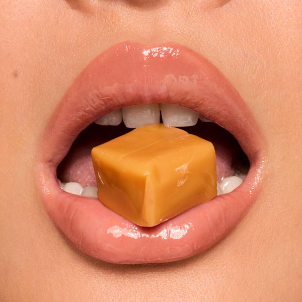 High-shine lips wear Caramel Latte, the ColourPop Fresh Kiss Glossy Balm, gripping a square caramel candy between the teeth in a tight macro close-up, highlighting the glossy finish, smooth texture, and warm nude sheen.