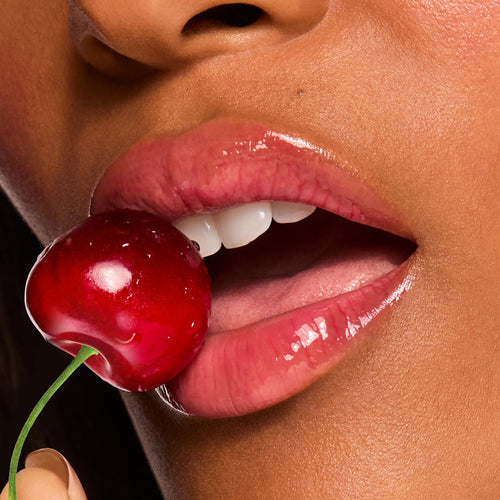 Glossy pink lips wear ColourPop Fresh Kiss Glossy Balm in Cherry Glaze, gripping a red cherry between the teeth in a macro beauty shot, showcasing a high-shine, juicy finish and hydrating lip balm texture.