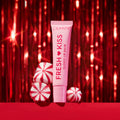 ColourPop Fresh Kiss Glossy Lip Balm in a pink squeeze tube stands upright beside red-and-white peppermint candies on a red surface, framed by sparkling red tinsel for a festive holiday beauty gift scene.