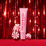 ColourPop Fresh Kiss Glossy Lip Balm in a pink squeeze tube stands upright beside red-and-white peppermint candies on a red surface, framed by sparkling red tinsel for a festive holiday beauty gift scene.