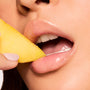 Glossy lips grip a fresh mango slice, showcasing sheer shine from Pretty in Pastels ColourPop Glossy Lip Balm Trio in a tight beauty close-up with natural skin texture, highlighting a hydrating, non-sticky finish.