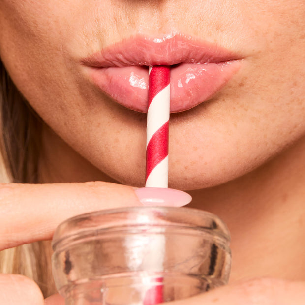 Glossy pink lips wearing ColourPop Fresh Kiss Glossy Balm in Strawberry Milk sip from a glass bottle with a red-and-white striped straw, showcasing high-shine hydration and a sheer milky tint.