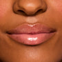 Glossy lips wearing ColourPop Fresh Kiss Glossy Balm in Vanilla Cake, showing a sheer nude-pink shine and smooth, hydrated finish in a close-up lip swatch on a model under studio lighting.