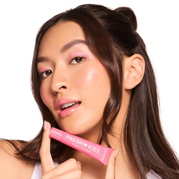 Model holds a pink ColourPop Fresh Kiss tube to glossy pink lips, showcasing Fruit Basket, the ColourPop Glossy Lip Balm Trio, in a close-up studio shot on white, emphasizing hydrating shine and a soft pink tint.