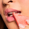 Glossy pink lips wearing Watermelon Sugar, the ColourPop Fresh Kiss Glossy Balm, softly bite a juicy watermelon slice held by manicured fingers, in a close-up studio beauty shot that showcases high-shine, hydrating color.
