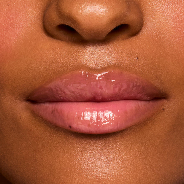 Glossy, hydrated lips wearing ColourPop Fresh Kiss Glossy Balm in Watermelon Sugar, showing a sheer pink shine on a close-up of the mouth against warm skin, highlighting cushiony texture and high-shine finish.