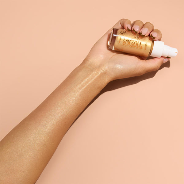 Hand holds a clear glass bottle with white pump of ColourPop SOL Glow Oil, It’s a Wolf Thing, while the golden oil is swatched along the forearm, revealing a luminous, sun-kissed sheen against a peach background.
