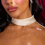 ColourPop Who-Hash Glitter Gel shimmering across a model’s collarbone and chest, scattering star-shaped sparkles; close-up with glossy lips, satin ivory choker, and rhinestone drop earrings against a deep red curtain backdrop.