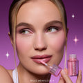 Model applies Gloss Me Up ultra glossy lip set in Heart to Get, holding the pink shimmery ColourPop-style tube against a purple starry background, showcasing high-shine nude-pink lips and matching soft glam makeup for a glossy finish.