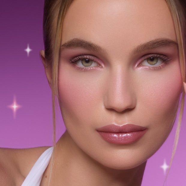 Model wearing Heart to Get All Eyes On Me Shadow Stix Set, showing shimmering pink eye shadow and soft blush on flawless skin against a glowing purple background with star accents, highlighting the eye makeup look for US beauty shoppers.