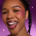 Smiling model wearing Heart to Get Sorry Not Sorry blush and UGL set, showing bold berry cheek color and glossy lips against a vibrant purple background with subtle sparkles.