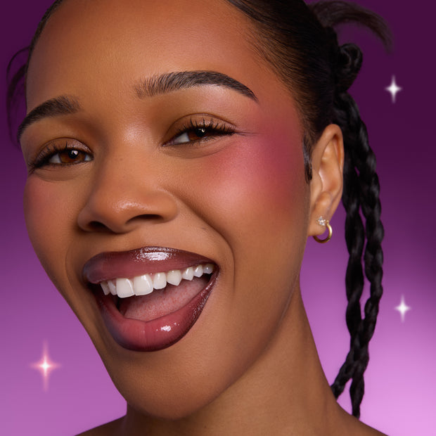 Smiling model wearing Heart to Get Sorry Not Sorry blush and UGL set, showing bold berry cheek color and glossy lips against a vibrant purple background with subtle sparkles.
