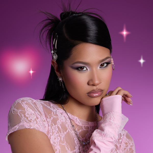 Model wearing shimmery lavender and silver eye look created with the Heart to Get shadow palette, posing against a glowing purple background with soft heart lights, showcasing bold graphic eyeshadow and glossy lips for Heart to Get makeup.