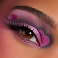 Close-up of a brown eye wearing the Heart to Get shadow palette, featuring bold pink and purple shimmer with heart-shaped accents at the inner and outer corners, showcasing dramatic, romantic eye makeup on warm-toned skin.