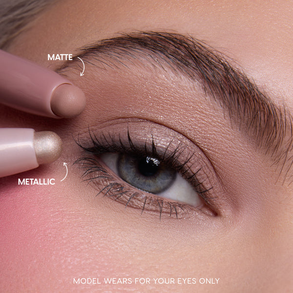 Close-up of a fair-skinned model’s blue eye wearing shimmery neutral shadow created with the All Eyes On Me Heart to Get Shadow Stix Set, showing matte and metallic cream eyeshadow tips applying blendable color on the eyelid.