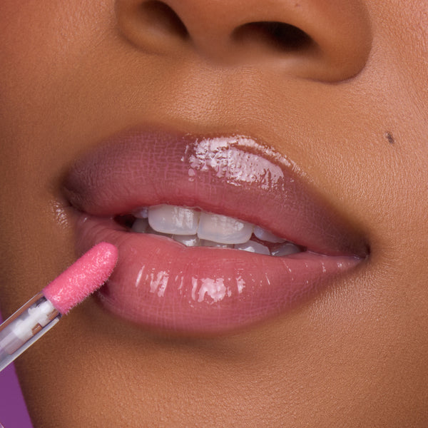 Close-up of a model’s lips applying Still That Girl Ultra Glossy Lip from Heart to Get, showing a sheer pink, high-shine gloss on smooth lips against a soft purple background for a glamorous beauty look.