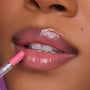 Close-up of a model’s lips applying Still That Girl Ultra Glossy Lip from Heart to Get, showing a sheer pink, high-shine gloss on smooth lips against a soft purple background for a glamorous beauty look.