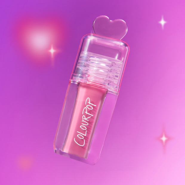 ColourPop Your New Obsession ultra glossy lip in the Heart to Get shade floats diagonally against a glowing pink and purple background, clear tube and heart-shaped cap highlighting the high-shine pink gloss for a dreamy makeup look.