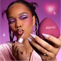 Model applies shimmery pink gloss while holding a heart-shaped ColourPop compact, showing bold purple and pink heart eye makeup that highlights the Heart to Get Full Collection full collection set against a dreamy pink background.