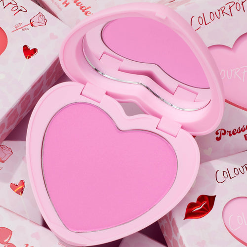 Heart-shaped Hot 2 Touch compact opens to reveal bright pink ColourPop Pressed Powder Blush, set in a pastel pink case with built-in mirror, resting atop Valentine-themed boxes with hearts and red lip graphics.