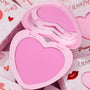 Heart-shaped Hot 2 Touch compact opens to reveal bright pink ColourPop Pressed Powder Blush, set in a pastel pink case with built-in mirror, resting atop Valentine-themed boxes with hearts and red lip graphics.