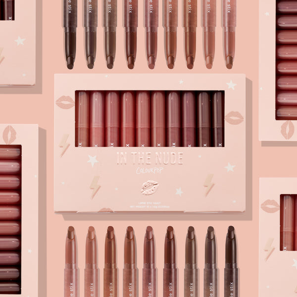 ColourPop Lippie Stix In the Nude vault displaying 10 nude shades - peach to deep cocoa - lined upright in a blush pink window box with star and lip icons, with additional Lippie Stix arranged around it on a soft pink backdrop.