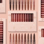 ColourPop Lippie Stix In the Nude vault displaying 10 nude shades - peach to deep cocoa - lined upright in a blush pink window box with star and lip icons, with additional Lippie Stix arranged around it on a soft pink backdrop.