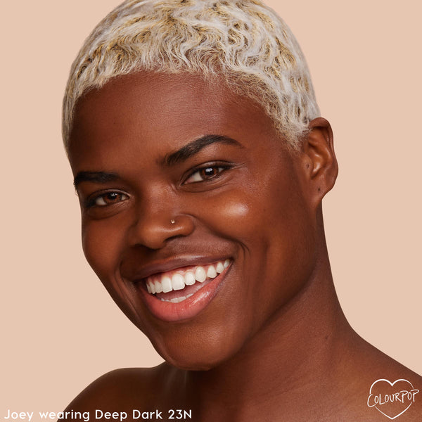 Model wearing Deep Dark 23 N smiles, showing smooth, radiant skin from the ColourPop Pretty Fresh Tinted Moisturizer; close-up face with short bleached waves on a warm beige studio background.