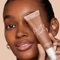 ColourPop Pretty Fresh Tinted Moisturizer, shade Deep Dark 21 W, held to a model’s face as the matte squeezy tube labeled Pretty Fresh and hyaluronic acid highlights a dewy, skin-like finish against a soft beige background.