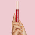 ColourPop Ultra Blotted Lip in Keep It Classy held upright by a hand with orange French tips, showcasing a clear tube of deep berry-red tint with a blush pink cap dotted with stars against a solid pink background.