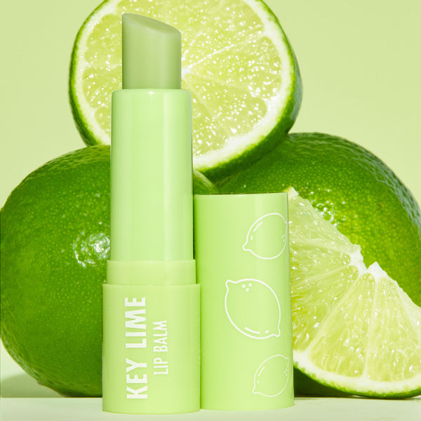 Key Lime ColourPop Lip Balm tube stands uncapped, balm bullet angled up, in glossy light‑green packaging with white “KEY LIME LIP BALM” text and citrus icons, set among whole and sliced limes on a pastel green backdrop.
