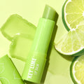 Key Lime ColourPop Lip Balm stick uncapped and twisted up, resting on a glossy swatch with the cap beside it, styled top-down on a lime-green background with fresh key lime wedges emphasizing the bright citrus flavor.