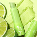 ColourPop Key Lime Lip Scrub stick uncapped and resting across two swipes of gritty green scrub, with sliced limes and a cap printed with citrus icons on a bright lime background, highlighting fresh, zesty exfoliation.