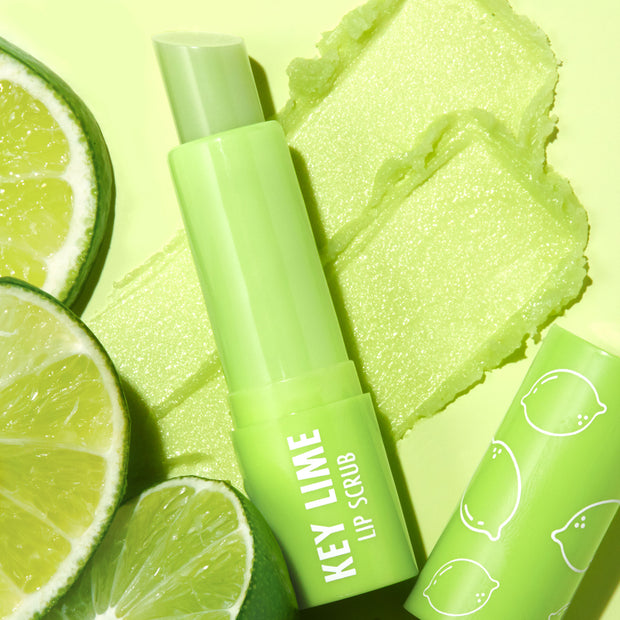 ColourPop Key Lime Lip Scrub stick uncapped and resting across two swipes of gritty green scrub, with sliced limes and a cap printed with citrus icons on a bright lime background, highlighting fresh, zesty exfoliation.