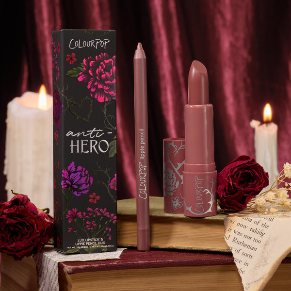 Anti-Hero ColourPop Lux Lipstick and matching lippie pencil stand uncapped beside the floral box, revealing a rosy-mauve bullet; displayed on vintage books with red roses and dripping candles against burgundy velvet for a moody, romantic vibe.