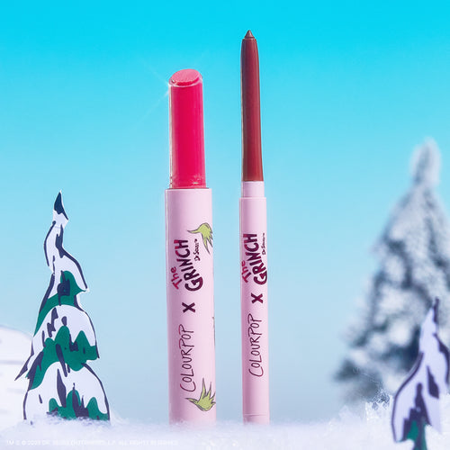 ColourPop Berry Grinchy So Juicy Plumping Gloss Stick stands uncapped with a vivid pink bullet beside a slim neutral lip pencil, both upright in snowy Grinch-themed scenery with fir trees and bright blue sky, collaboration branding on tubes.