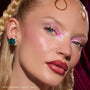 ColourPop Change of Heart Super Shock Highlighter Kit highlighting model’s cheekbones and inner corners in an icy pink glow, shown in a studio close-up with glossy skin, braided blonde hair, teal bow earrings, and a magenta draped backdrop.