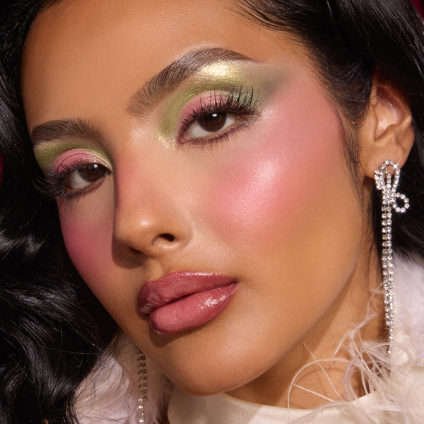 Model showcases shimmery green and pink eyeshadow, luminous blush, and glossy mauve lips using the ColourPop X The Grinch Full Collection Set by Colourpop, paired with long lashes, rhinestone bow earrings, and a feather-trim collar in a studio close-up.