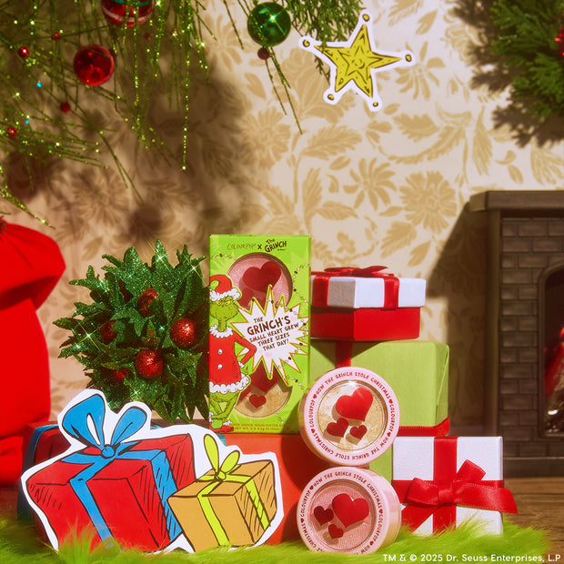 ColourPop x The Grinch holiday makeup set arranged as stacked gifts, featuring a green box with Grinch art and heart-shaped cutouts plus two round pots with red heart windows, surrounded by wrapped presents, ornaments, and a fireplace.